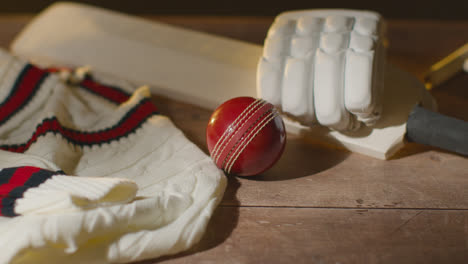 Cricket-Still-Life-With-Close-Up-Of-Bat-Ball-Gloves-Stumps-Jumper-And-Bails-Lying-On-Wooden-Surface-In-Locker-Room