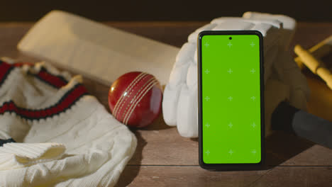 Mobiltelefon-Mit-Grünem-Bildschirm,-Umgeben-Von-Cricketschlägerball-Und-Kleidung-Auf-Holzoberfläche
