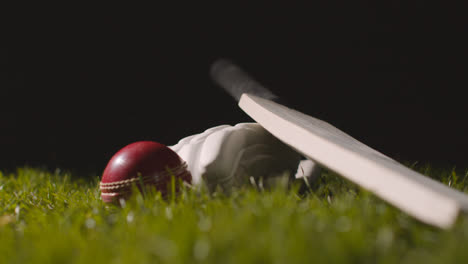 Studio-Cricket-Still-Life-With-Close-Up-Of-Ball-Rolling-Onto-Bat-And-Glove-Lying-In-Grass
