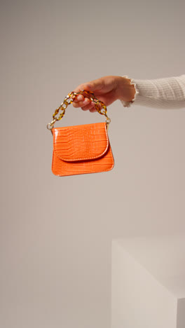 Close-Up-Vertical-Video-Of-Female-Social-Media-Influencer-Producing-User-Generated-Content-Holding-Out-Orange-Fashion-Handbag-1