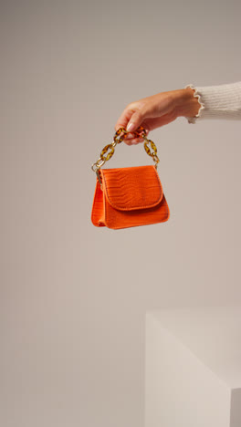 Close-Up-Vertical-Video-Of-Female-Social-Media-Influencer-Producing-User-Generated-Content-Holding-Out-Orange-Fashion-Handbag