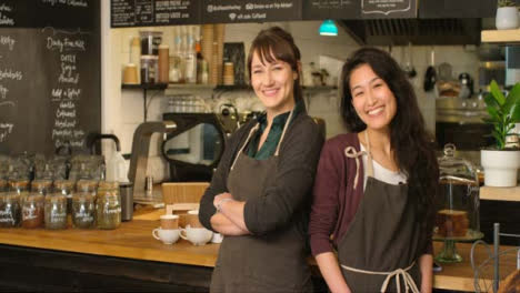 Portrait-of-two-happy-female-baristas-at-cafe-smiling-to-camera