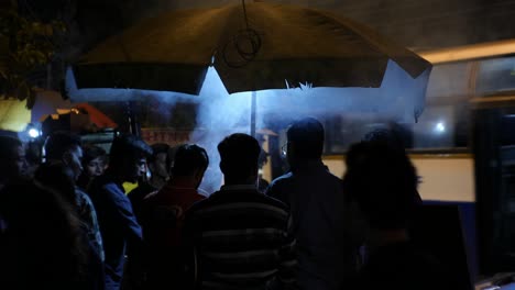 Bengaluru-Karnataka--India--January-25-2020-Medium-shot-of-people-waiting-under-an-umbrella-at-a-street-side-barbecue-shop-with-heavy-smoke-during-night-time