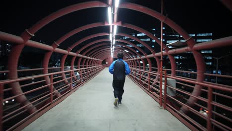 Wide-angle-follow-shot-of-a-man-jogging-on-a-metal-red-colored-skywalk-during-early-morning-hours