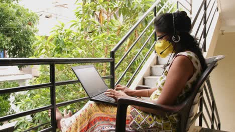 Closeup-of-an-Indian-woman-wearing-safety-mask-and-headset-microphone--working-with-a-laptop-sitting-in-the-balcony-during-the-Covid19-corona-virus-pandemic