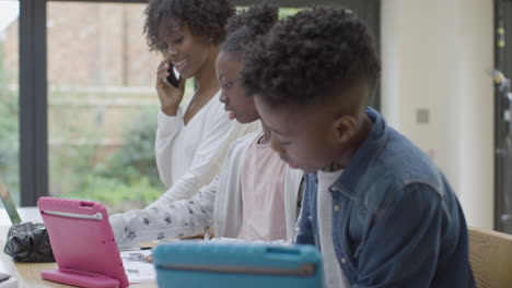 Mother-Helping-Two-Young-Children-with-Homework-On-Tablets-While-She-Works