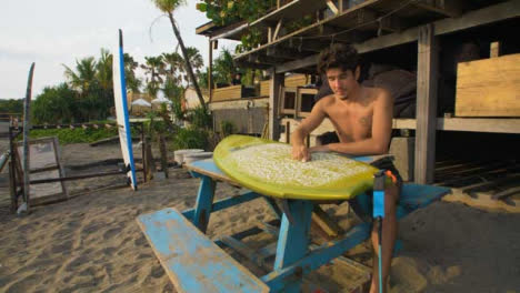 Tracking-Shot-Pulling-Away-from-Young-Man-Preparing-Surf-Board