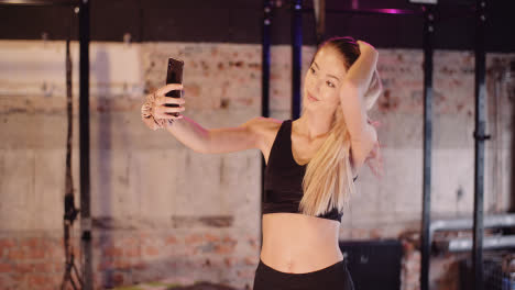 Hermosa-Atleta-Femenina-Ajustando-El-Cabello-Mientras-Toma-Selfie-A-Través-Del-Teléfono-Inteligente-En-El-Estudio-De-Salud