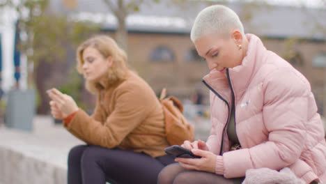 Mittlere-Aufnahme-Von-Zwei-Jungen-Frauen,-Die-Draußen-Auf-Ihren-Handys-Sitzen