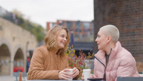 Low-Angle-Shot-of-Two-Female-Friends-Sitting-at-Outdoor-Table-Drinking-Coffee