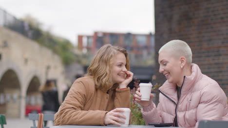 Low-Angle-Shot-of-Two-Young-Women-Sitting-at-Outdoor-Table-Drinking-Some-Coffee