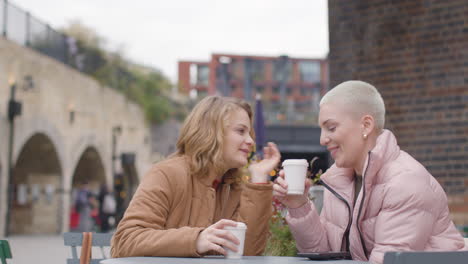 Low-Angle-Shot-of-Two-Young-Women-Sitting-at-Outdoor-Table-Drinking-Cups-of-Coffee