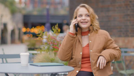 Plano-Medio-De-Una-Mujer-Joven-Sentada-En-Una-Mesa-Al-Aire-Libre-Respondiendo-Llamadas-Telefónicas