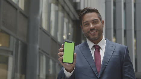 Portrait-Of-Businessman-Outside-City-Of-London-Offices-Holding-Up-Green-Screen-Mobile-Phone