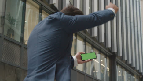 Excited-Businessman-Outside-Office-Holding-Green-Screen-Mobile-Phone-Celebrating