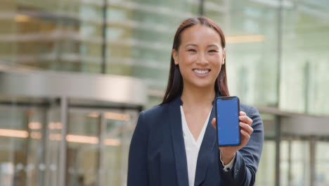 Portrait-Of-Businesswoman-Outside-City-Of-London-Offices-Holding-Blue-Screen-Mobile-Phone