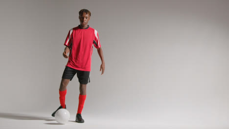 Studio-Portrait-Of-Young-Male-Footballer-Wearing-Club-Kit-Kicking-Ball-Towards-Camera
