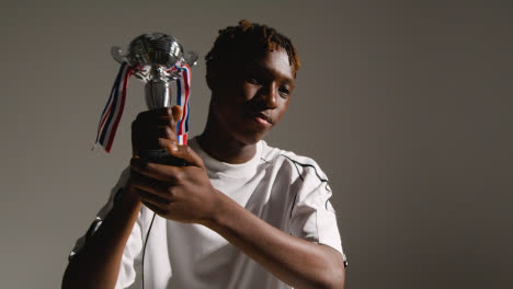 Studio-Portrait-Of-Young-Male-Footballer-Or-Sportsperson-Wearing-Club-Kit-Celebrating-Holding-Trophy