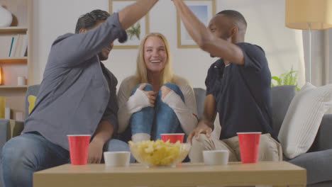 Multi-Cultural-Friends-Celebrating-Watching-Sports-Game-On-TV-Sitting-On-Sofa-At-Home-Eating-Chips-And-Dips-