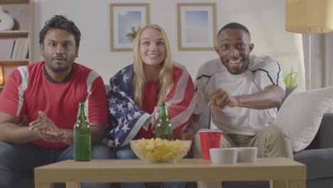 Multi-Cultural-Friends-Supporting-USA-Celebrating-Watching-Sports-Game-On-TV-Sitting-On-Sofa-At-Home-With-Flag