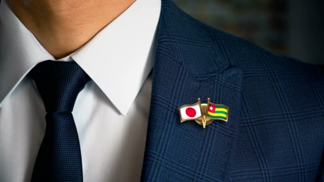 Businessman-Walking-Towards-Camera-With-Friend-Country-Flags-Pin-Japan---Togo