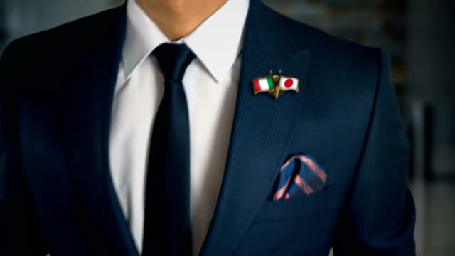 Businessman-Walking-Towards-Camera-With-Friend-Country-Flags-Pin-Italy---Japan