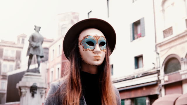 Woman-with-long-hair-and-hat-wearing-a-white-carnival-face-mask-in-Venice-street,-Italy-looking-at-camera,-turning-back.