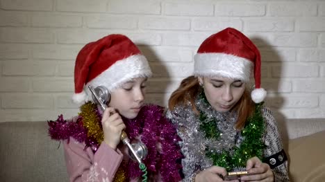 Two-funny-little-girls-in-Santa's-hats-are-calling-on-the-phone.-They-sit-on-the-couch-with-Christmas-tinsel-on-their-shoulders,-dance-and-sing-into-the-phone-against-the-white-wall.