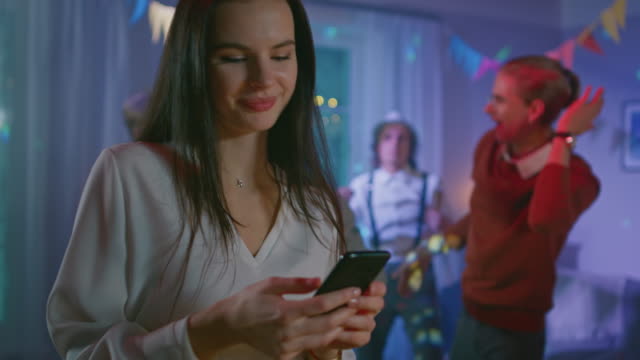 At-the-Wild-House-Party:-Beautiful-Young-Girl-Uses-Smartphone,-Browses-Through-Internet-and-Social-Network.-In-the-Background-Crowd-of-Young-People-Dancing-Off-and-Having-Fun.