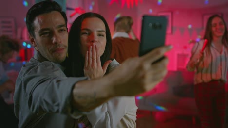 At-the-Wild-House-Party:-Beautiful-Young-Couple-Take-Selfies-with-a-Smartphone-for-Social-Network-Sharing.-In-the-Background-Crowd-of-Young-People-Dancing-Off-and-Having-Fun.