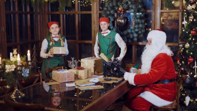 Elves-in-green-suits-and-red-hats-is-dancing-with-gifts-in-their-hands-and-putting-it-on-the-table-with-letters-of-congratulations-and-a-magnifying-glass-near-Santa-Claus-on-the-background-of-the-Christmas-tree-and-New-Year-decorations-in-the-room