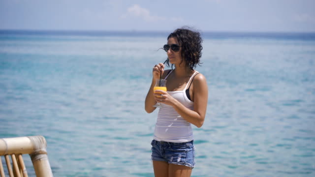 Ein-junges-Mädchen,-bekleidet-mit-einem-weißen-T-shirt-und-kurzen-Jeansshorts-stehen-neben-dem-blauen-Meer-und-der-Spaß-ist-Orangensaft-durch-einen-Strohhalm-trinken.