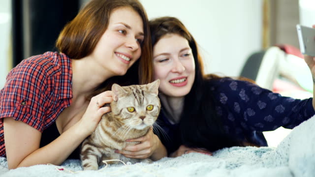 Zwei-glückliche-Frauen-Freunde-im-Bett-liegen-und-dass-Selfie-mit-Katze-und-viel-Spaß-im-Bett-zu-Hause