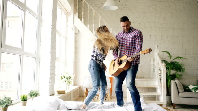 Funny-happy-and-loving-couple-dance-on-bed-singing-with-tv-controller-and-playing-guitar.-Man-and-woman-have-fun-during-their-holiday-at-home