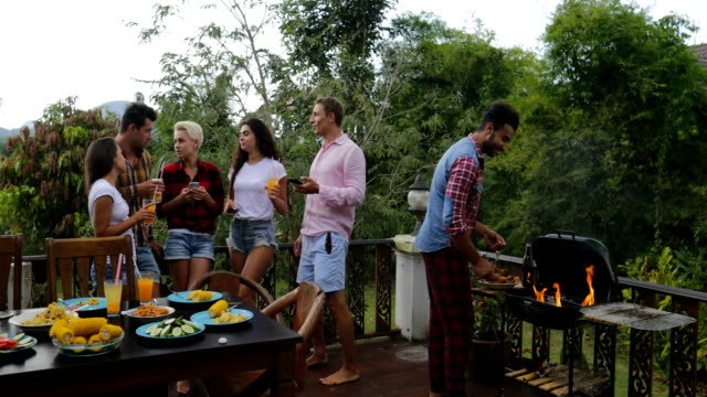 Young-People-Talking-Using-Cell-Smart-Phones-While-Latin-Man-Cooking-Barbecue-Friends-Group-Gathering-On-Summer-Terrace-Communication