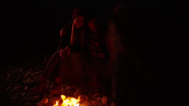 Young-couple-sitting-around-campfire-at-the-beach-at-night