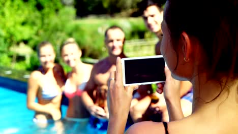 Mujer-sonriente-haciendo-clic-fotos-de-amigos-de-teléfono-móvil-junto-a-la-piscina