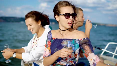two-young-pretty-brunettes-are-drinking-champagne-on-a-pleasure-boat-in-summer-day,-looking-and-showing-at-a-sea