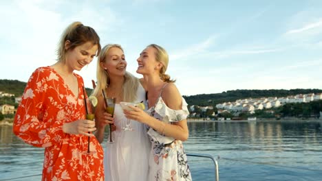 Tres-hermosas-mujeres-jóvenes-con-vestidos-de-verano-beber-cócteles-en-un-yate,-reír-y-pasarlo-bien.-Estado-del-mar-y-pueblos-de-isla-pequeña-en-el-fondo.
