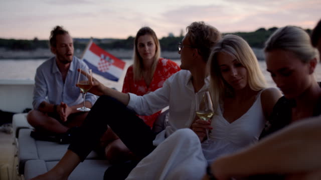 Group-of-Young-People-Talk,-Drink-Champagne-in-the-Stern-of-the-Moving-Yacht.-They-Have-Great-Vacation.-In-the-Background-Island-with-Small-Village.
