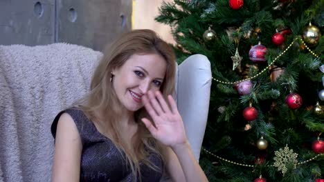 A-beautiful-smiling-woman-in-a-shiny-dress-waving-and-sitting-on-a-chair-near-a-christmas-tree