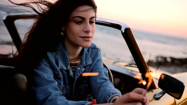 Woman-in-convertible-car-holding-sparklers-at-beach-at-sunset