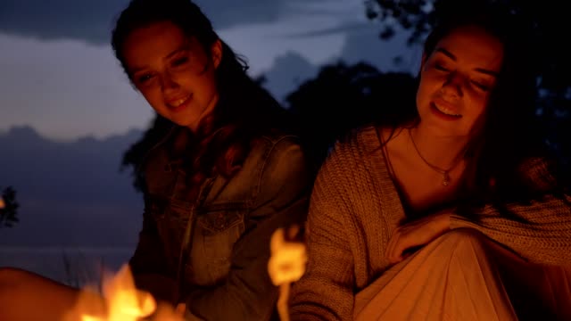 Zwei-junge-Kaukasische-Mädchen-sitzen-abends-am-Feuer-in-der-Natur,-Marshmelow,-Kochen,-lachen,-Essen