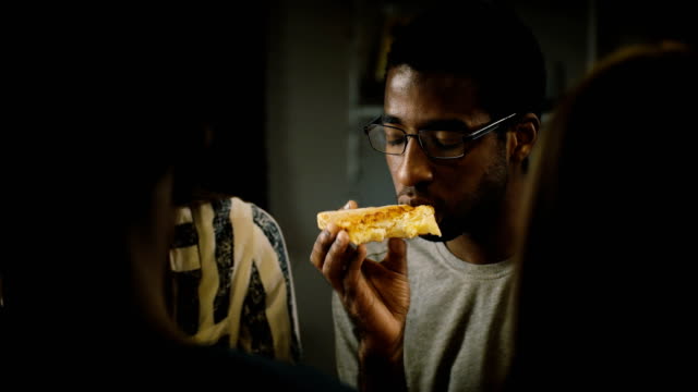 Pizza-de-afroamericano-joven-comiendo-en-una-fiesta-informal.-Los-jóvenes-disfrutan-de-comida-rápida-en-la-cocina-en-casa