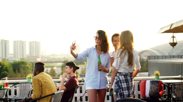 Fröhliche-Freundinnen-nehmen-Selfie-mit-Smartphone-und-lachen-stehen-auf-Dach-mit-Getränke-in-Flaschen,-die-Partei-zu-genießen.-Technologie,-Getränke--und-Jugend-Konzept.