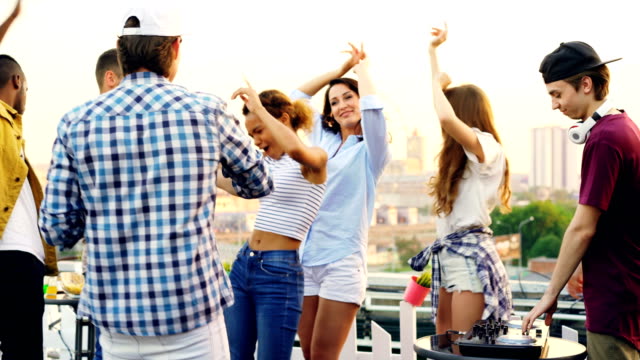 Young-people-are-dancing-and-laughing-while-male-DJ-is-working-with-equipment-at-rooftop-party-on-summer-day.-Entertainment,-youth-and-music-concept.