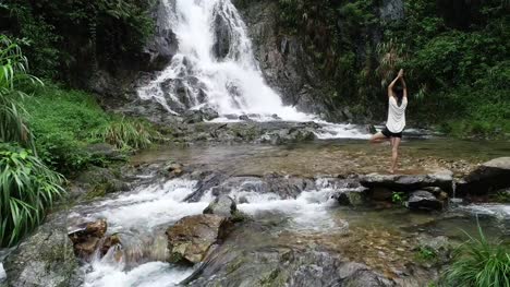 Vista-aérea-de-mujer-de-estilo-de-vida-saludable-haciendo-yoga-cerca-de-cascada-en-el-bosque