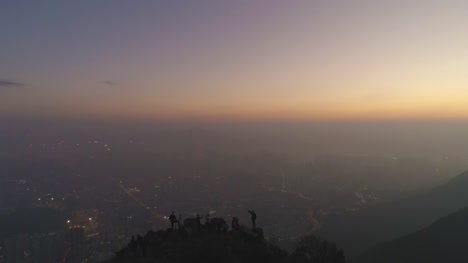 Silhouettes-of-people-on-the-top-of-rock-at-twilight.-City-Skyline-in-Smog.-Aerial-View.-Drone-is-Flying-Upward.-Establishing-shot.