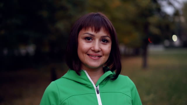 Portrait-of-pretty-young-woman-in-sportswear-looking-at-camera-and-smiling-standing-outdoors-in-park-in-autumn-evening-with-lights-visible-in-background.