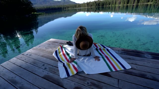Woman-sitting-on-wooden-pier-by-stunning-alpine-lake-contemplating-nature